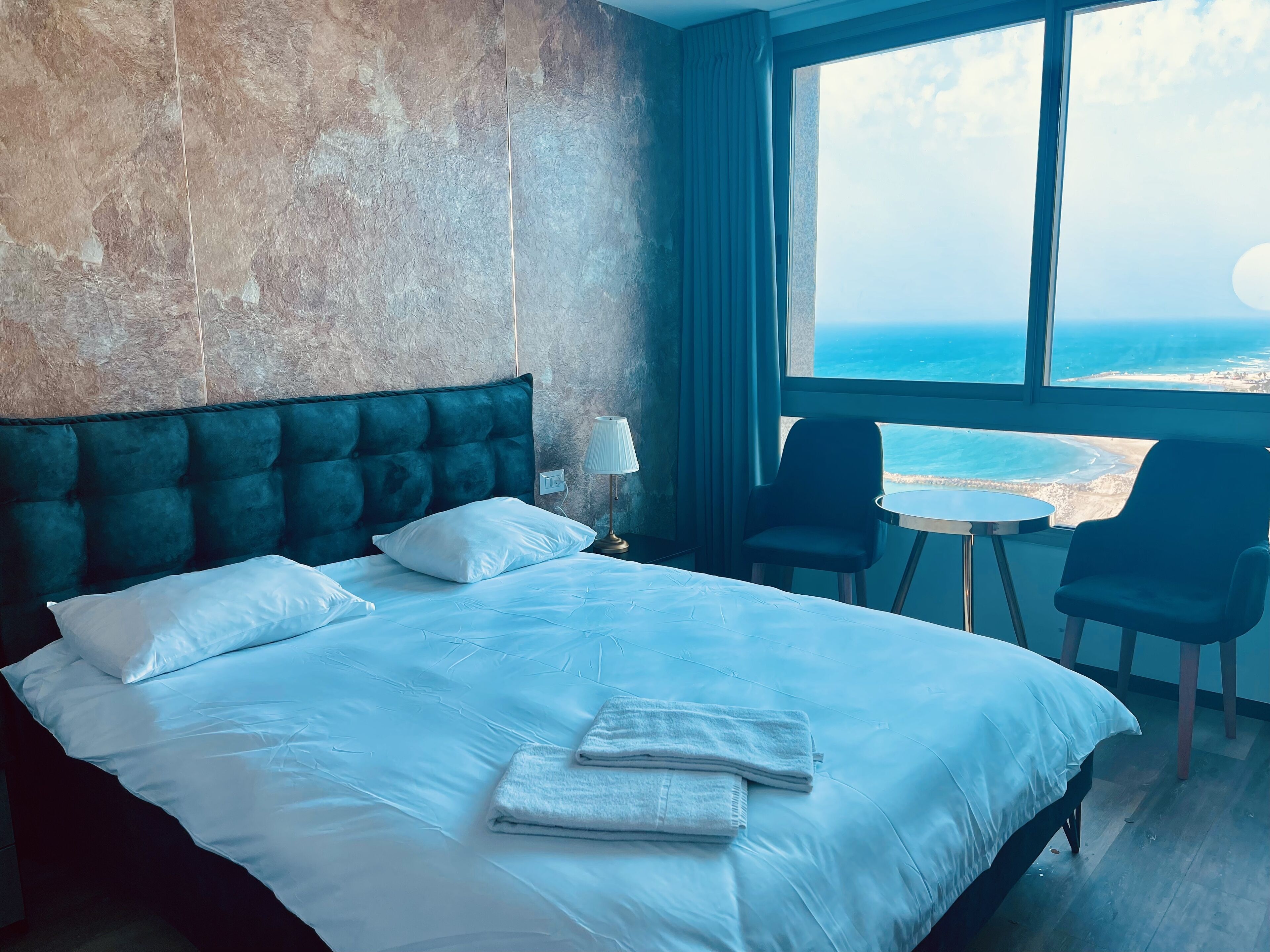 A hotel room with a large bed, a view of the sea, a small table, and two chairs.