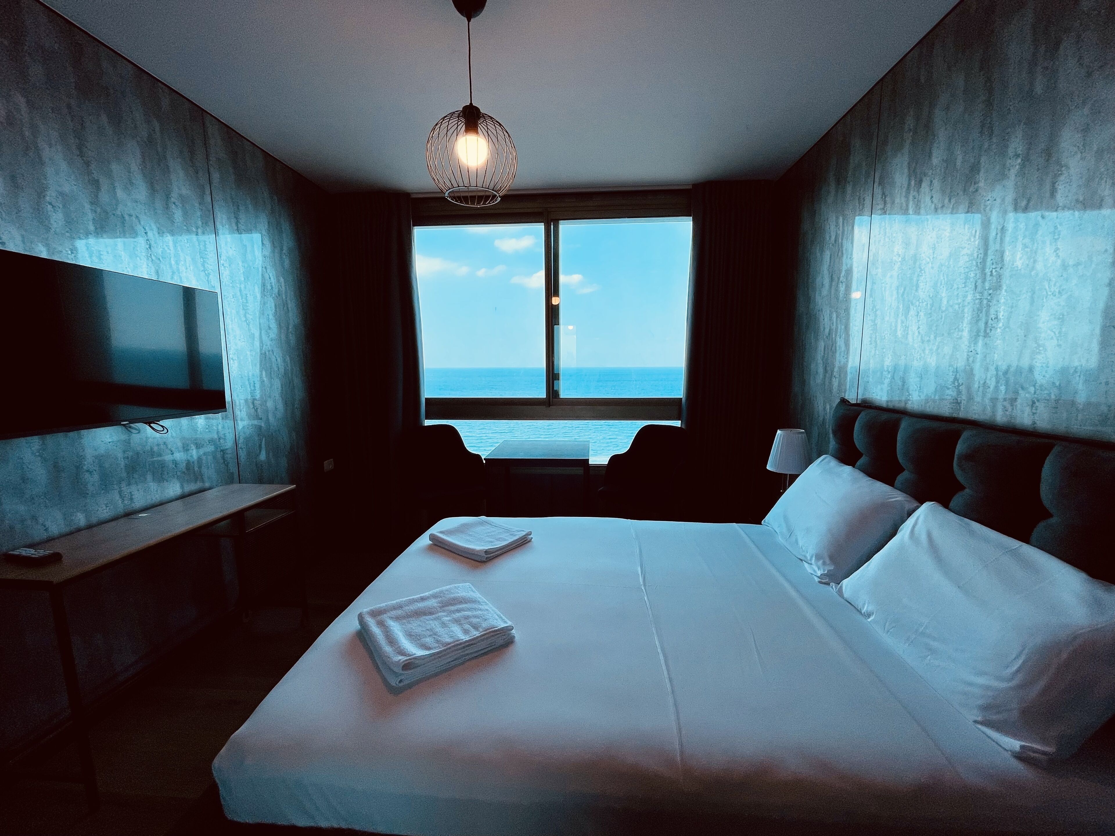 A hotel room with a large bed, a flat-screen TV, a window with a view of the sea, and a hanging light fixture.