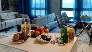 A modern living room with a sofa, a dining table with breakfast items, and a view of the ocean through large windows. - Carmel Holiday Apartments (Netanya)