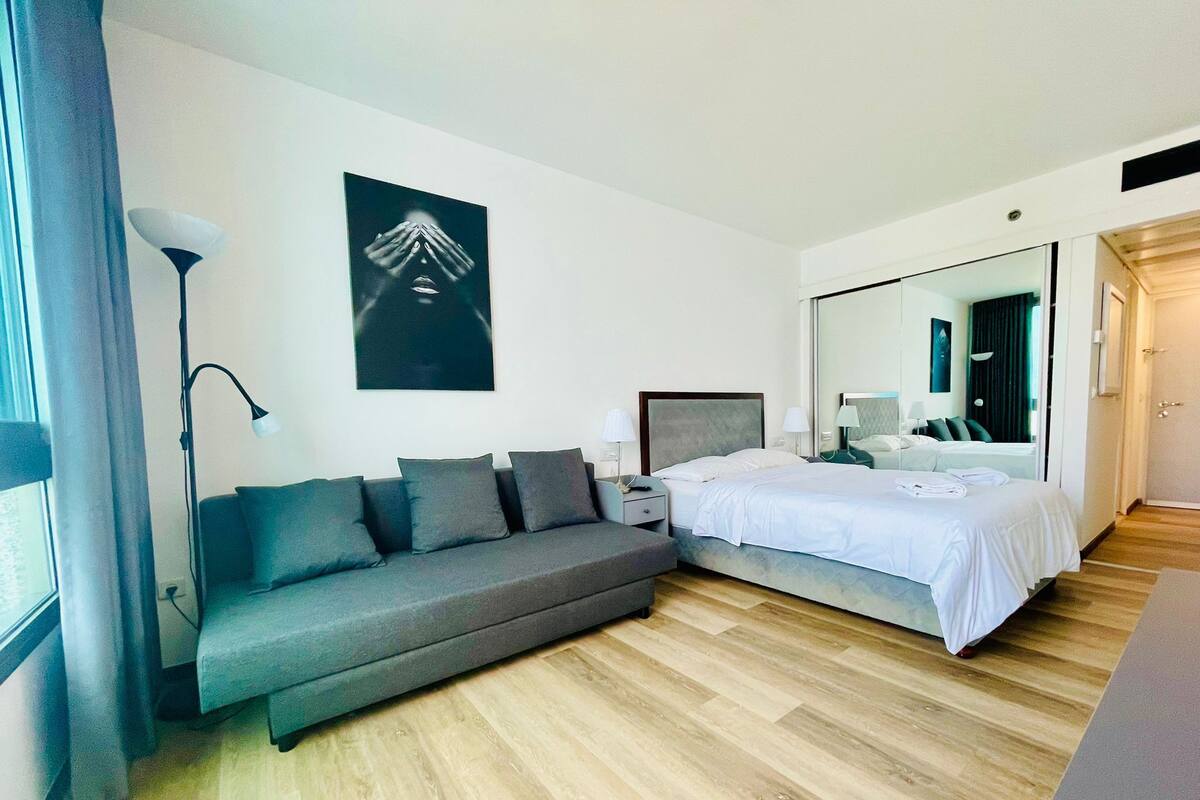 A modern bedroom with a grey sofa, a large bed with white linens, a bedside table with a lamp, and a large mirror.