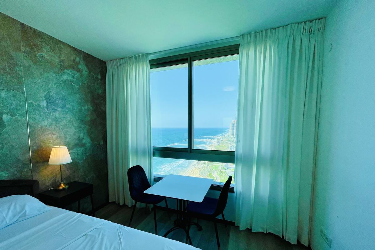 A hotel room with a bed, a desk, a chair, a lamp, and a window with curtains.