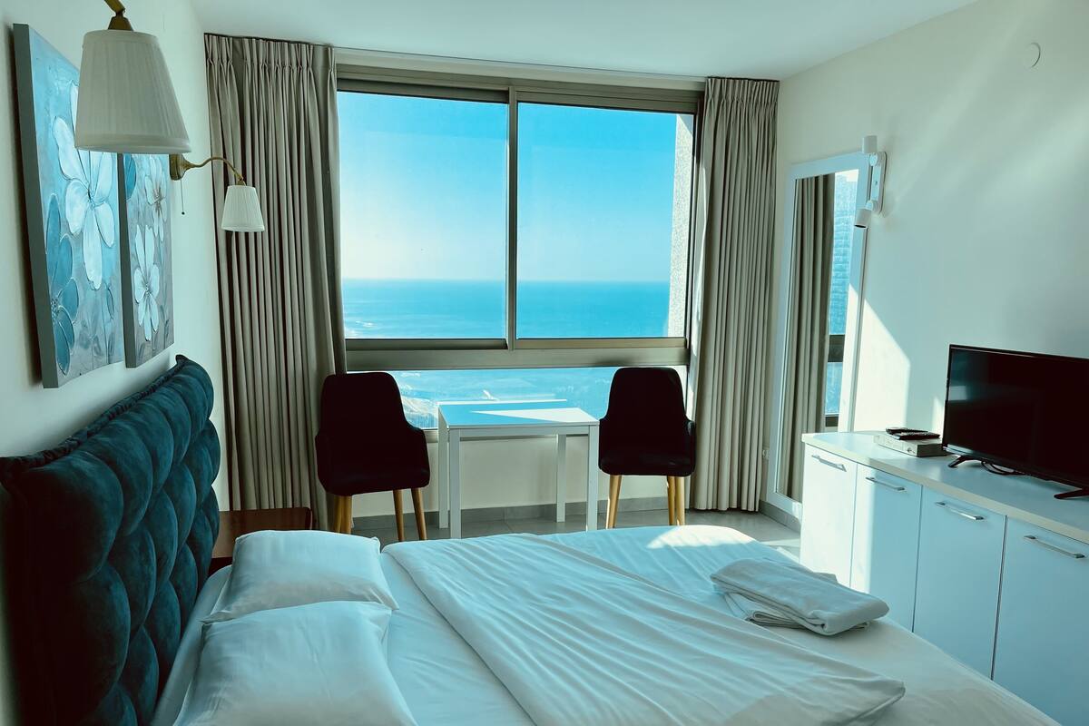 A hotel room with a bed, a desk, two chairs, a TV, and a large window with a view of the ocean.