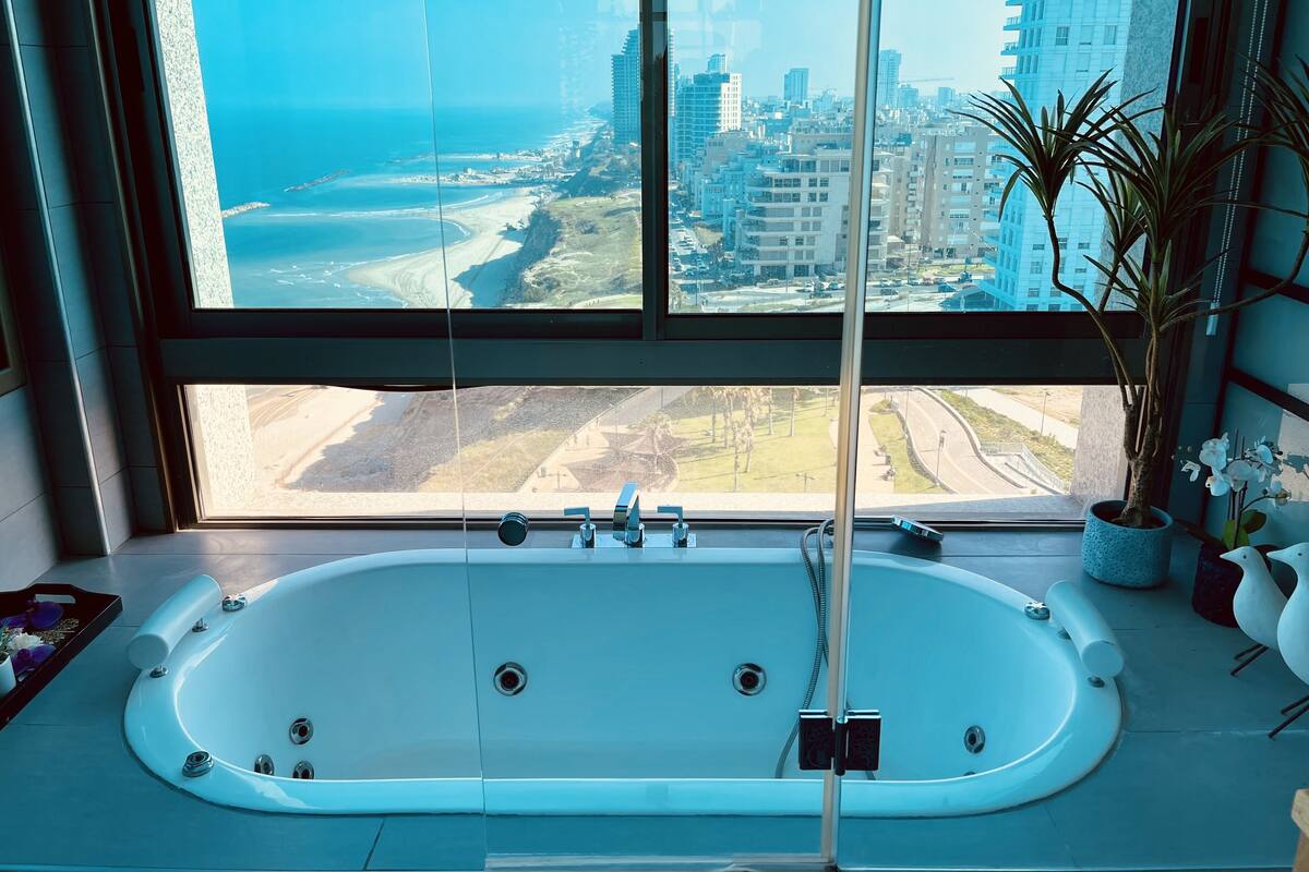 A modern bathroom with a large bathtub, a window with a city view, and a potted plant.