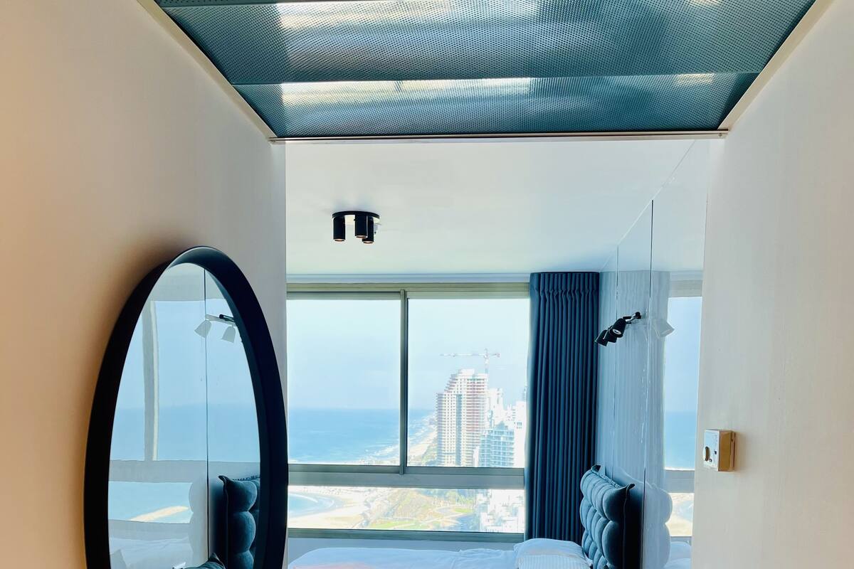 A modern hotel room with a large window, a round mirror, a bed, a sofa, and a ceiling light.