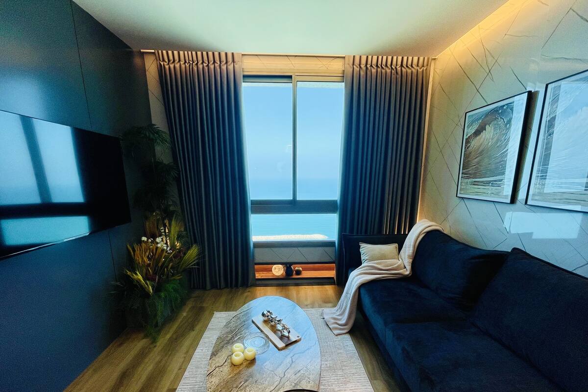 A modern living room with a dark blue sofa, a wooden coffee table, a flat-screen TV, and a large window with curtains.