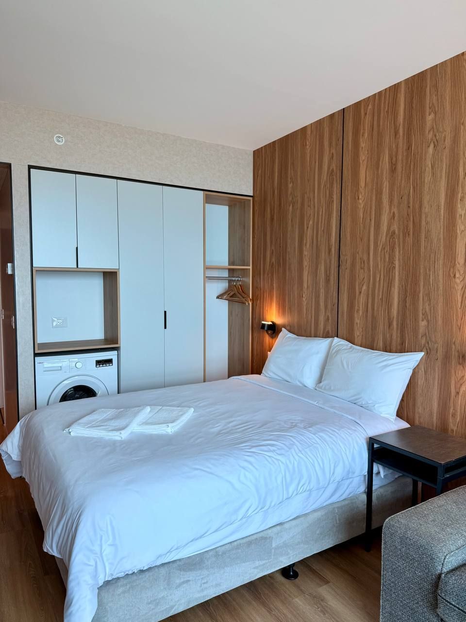 Un dormitorio moderno con una cama grande, lavadora integrada, paneles de madera y una mesita de noche pequeña.