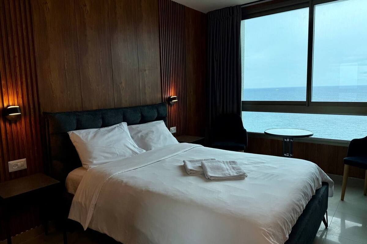 A hotel room with a large bed, a nightstand, a round table, and a view of the sea.