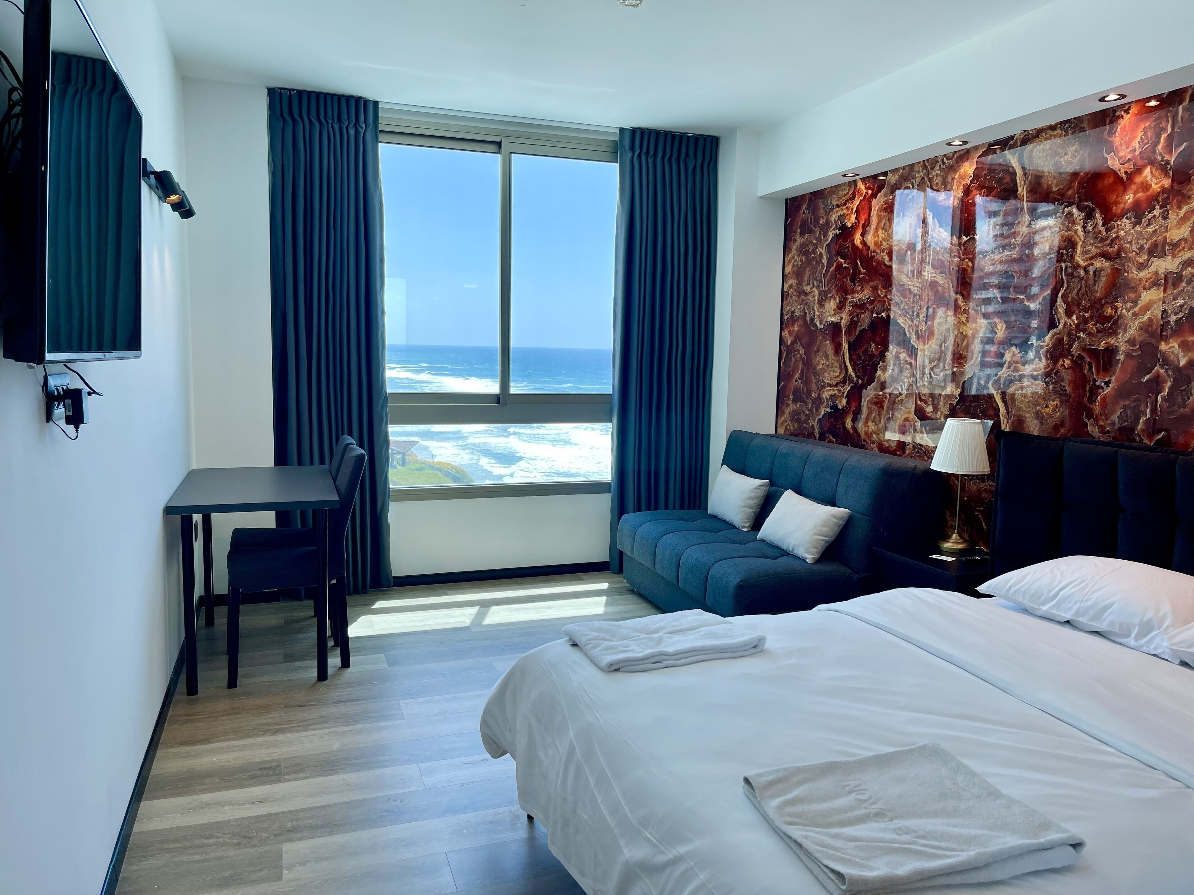 Ein Hotelzimmer mit einem Bett, einem Schreibtisch, einem Sessel und einem Sofa, mit einem großen Fenster und Blick auf den Ozean.