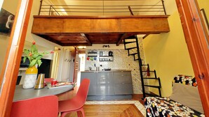 A compact kitchen with a built-in stove, a dining area with a red chair, and a lofted sleeping area with a patterned bed. - 4BNB - Cozy Vatican Loft with Terrace (Roma)