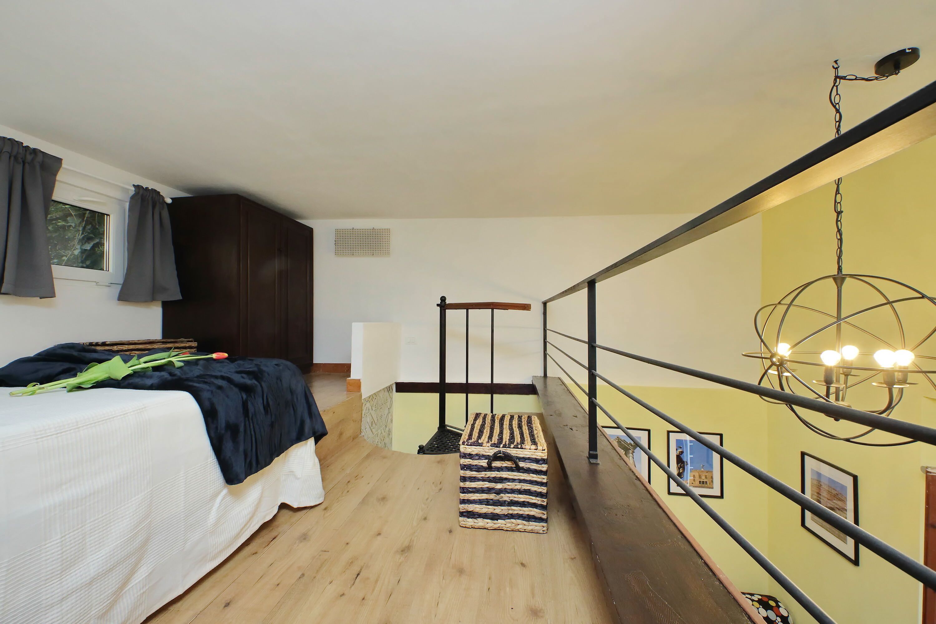 A bedroom with a bed, a wooden floor, a black and white striped storage box, a black metal railing, and a yellow wall with framed pictures.