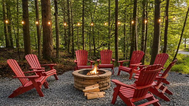 A fire pit surrounded by red Adirondack chairs in a forest setting.