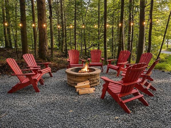 A fire pit surrounded by red Adirondack chairs in a forest setting.