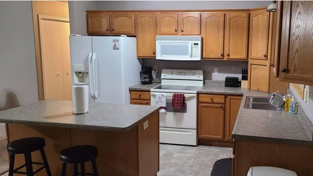 A compact kitchen with wooden cabinets, a white refrigerator, a microwave, a stove, and a sink. There are two stools at the counter. - 5 Bedroom Home for your Black Hills Adventures (Rapid City)