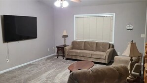 A living room with a flat-screen TV, a sofa, a coffee table, a lamp, and a window with blinds. - 5 Bedroom Home for your Black Hills Adventures (Rapid City)