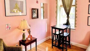 Una habitación con paredes rosas, piso de madera, una pequeña mesa con lámpara, dos sillas y obras de arte enmarcadas.
