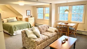 A bedroom with a bed, a sofa, a dining table with chairs, and a window with a view of trees.