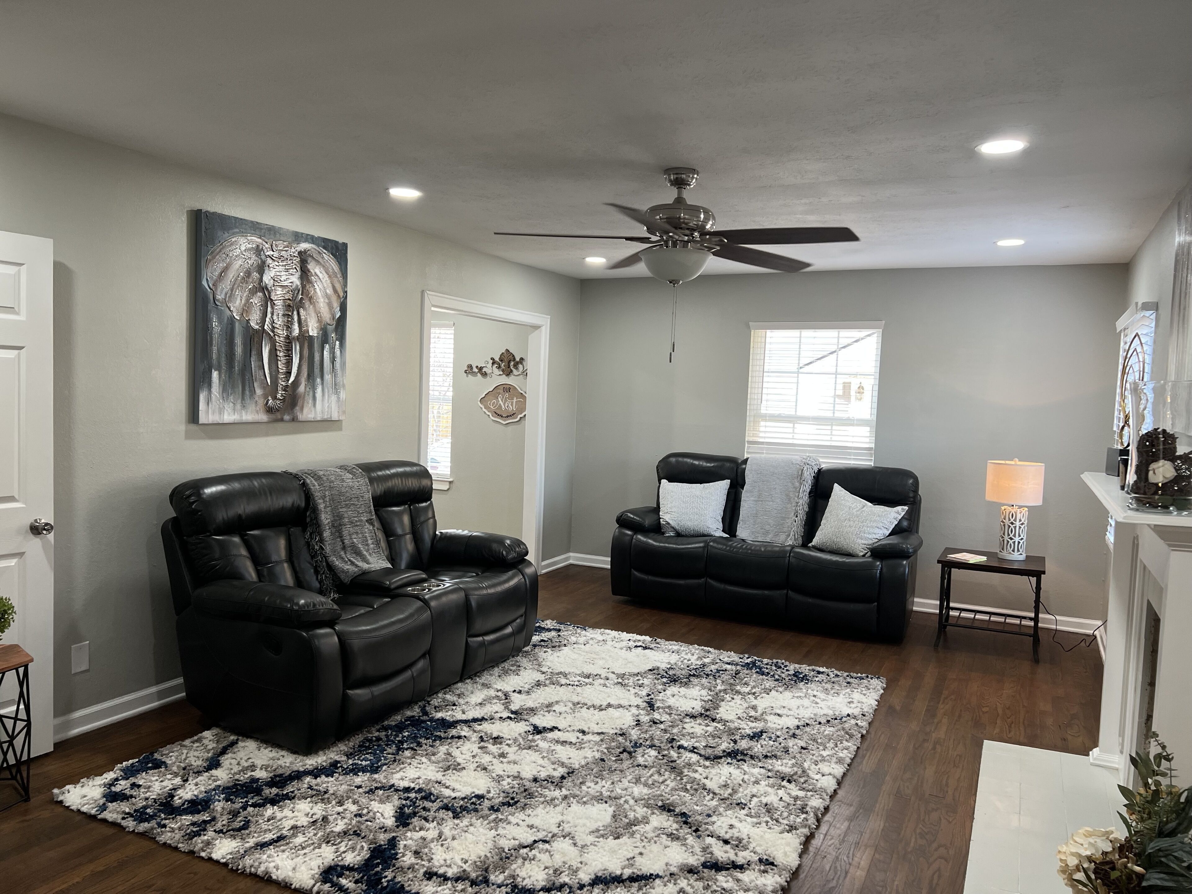 A living room with a black leather sofa, a patterned rug, a ceiling fan, and a window with blinds.