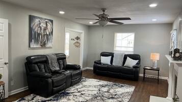 A living room with a black leather sofa, a patterned rug, a ceiling fan, and a window with blinds.