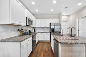A modern kitchen with white cabinets, granite countertops, stainless steel appliances, and a wooden floor. - Modern 3BR Furnished Home Near DIA with 2-Car Garage (Denver)