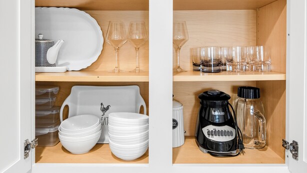 A kitchen cabinet with shelves containing white plates, bowls, wine glasses, and a coffee maker.