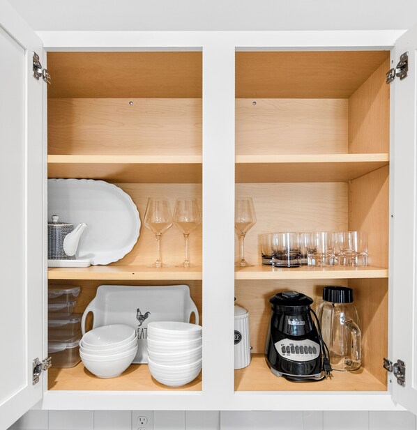 A kitchen cabinet with shelves containing white plates, bowls, wine glasses, and a coffee maker.