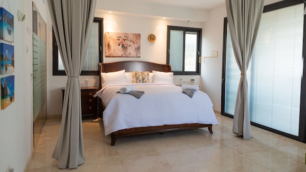 A bedroom with a bed, curtains, a nightstand, a painting, and a window. - Grace Holon Villa & Pool (Holon)