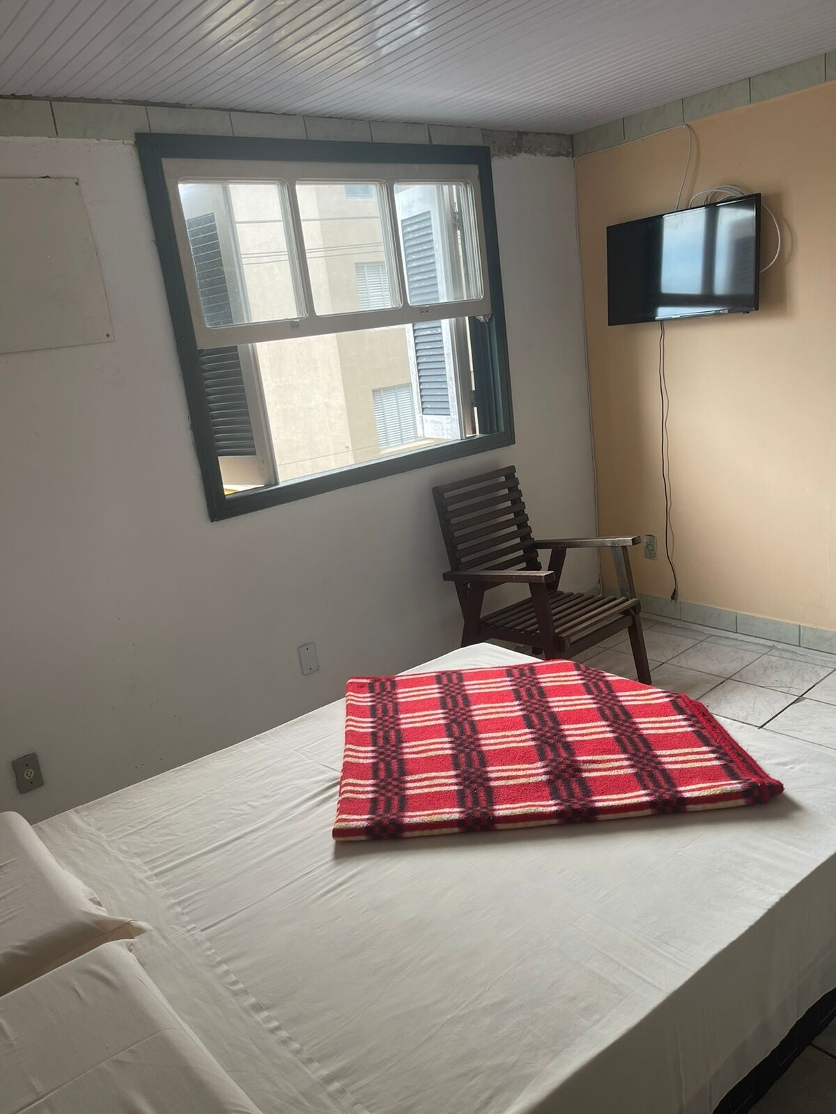 A room with a bed, a chair, a television mounted on the wall, and a window with blinds.
