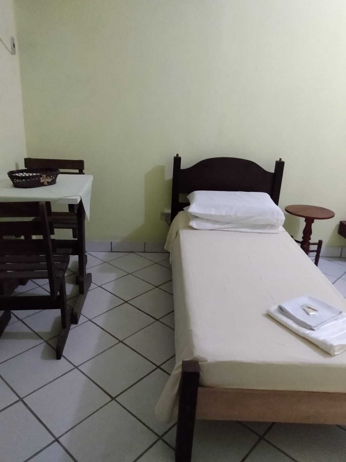 A single bed with a wooden frame and white bedding, a small table with a basket, and a tiled floor.