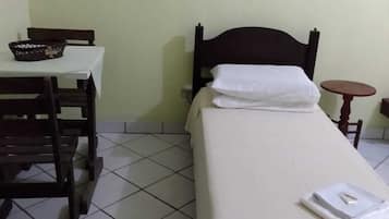 A single bed with a wooden frame and white bedding, a small table with a basket, and a tiled floor.