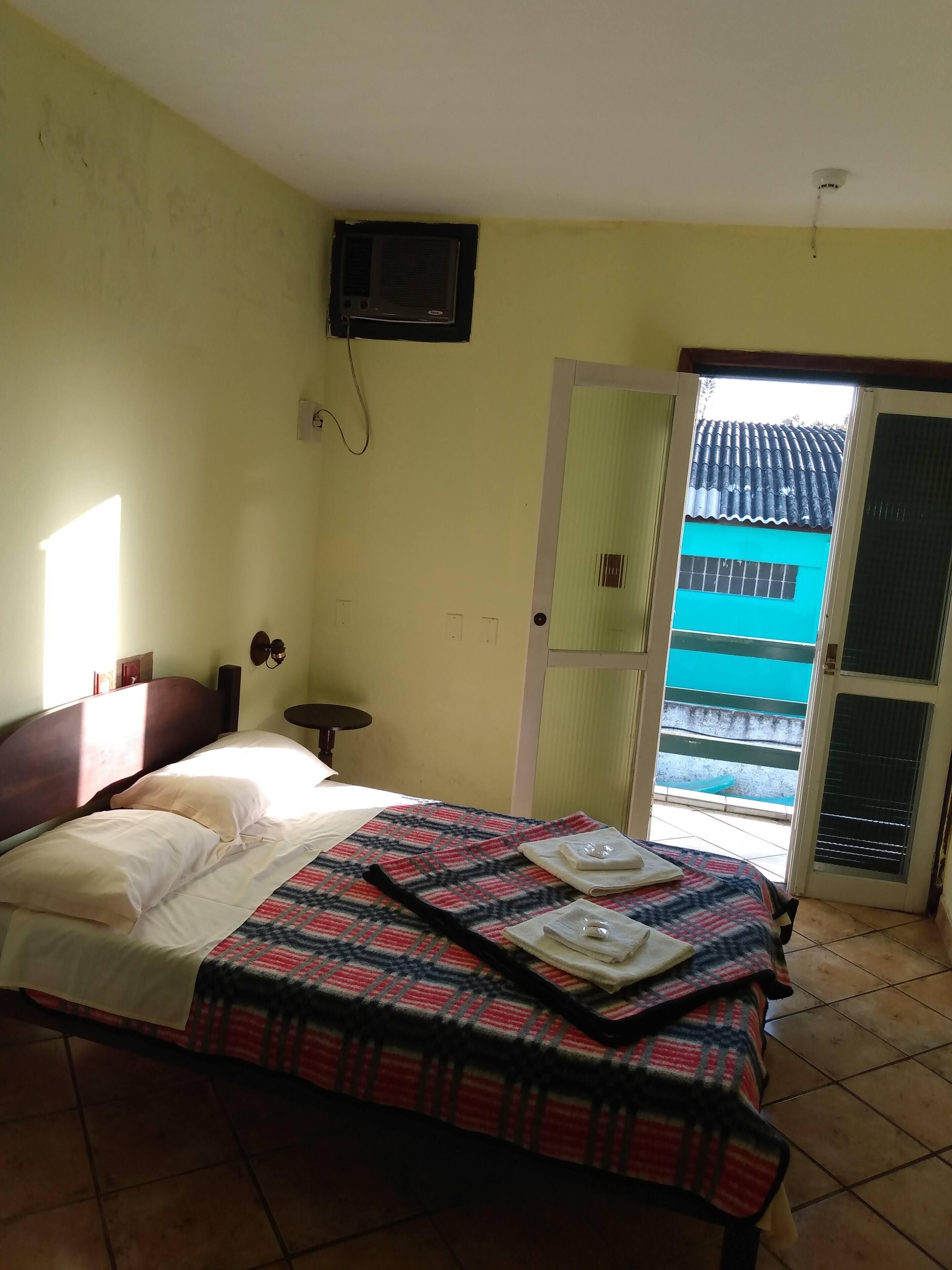 A hotel room with a bed, a television mounted on the wall, a small table, and a view of a blue building through an open door.