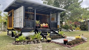 A modern house with a covered porch, a small outdoor seating area, and a garden with various plants and rocks. - Are Mii , a stylish one room container home (Arorangi District)