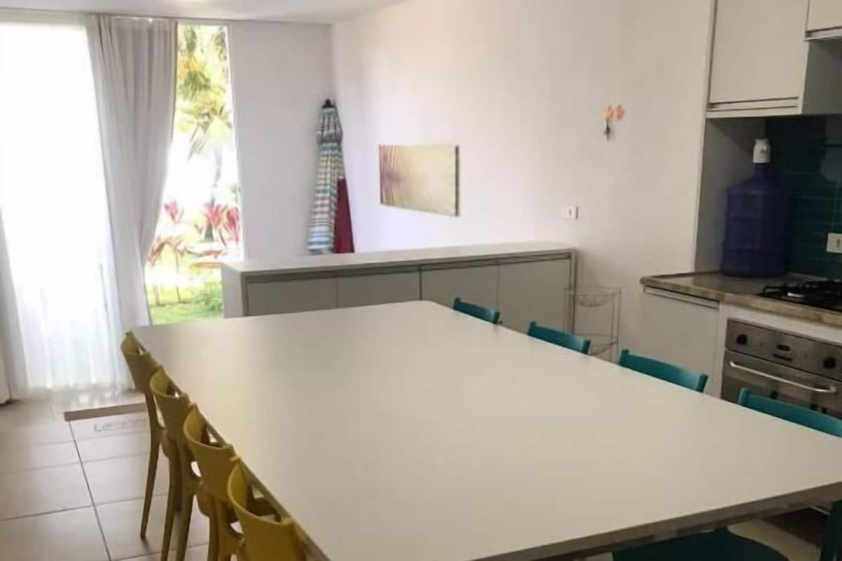 Un comedor con una mesa y sillas amplias, una cocina con armarios y dispensador de agua, y vista a un jardín a través de una ventana.