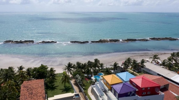 A beach view with a clear sky, palm trees, and a row of colorful buildings with a swimming pool. - Carneiros Flat Praia (Tamandaré)