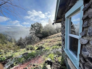 Un edificio de piedra con una ventana, un muro de piedra y vistas a una ladera boscosa.