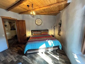 Une chambre avec un plafond en bois, un lit recouvert d’une literie à motifs, un luminaire fixé au mur et une porte donnant sur une autre pièce.