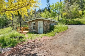 Eine kleine Steinhütte mit einer Holzterrasse und einer Tür, umgeben von Bäumen und einem Kiesweg.