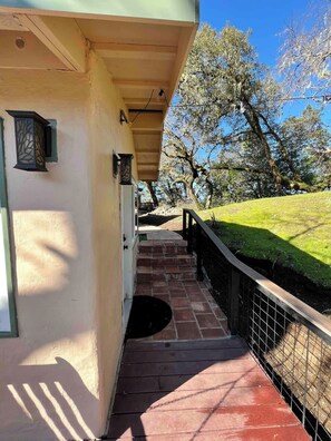 A covered outdoor area with a brick pathway leading to a door, surrounded by trees and a grassy slope.