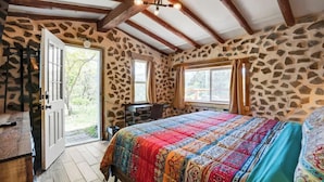 Une chambre avec un mur en pierre, un plafond en bois, un lit recouvert d’une courtepointe colorée et un bureau avec une chaise.