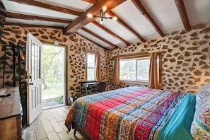 A bedroom with a stone wall, a wooden ceiling, a bed with a colorful quilt, and a desk with a chair.