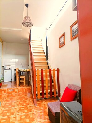 Un escalier avec une rampe en bois menant à un deuxième étage, un coin repas avec une table et des chaises, et une petite cuisine en arrière-plan.