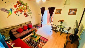 Ein Wohnzimmer mit einer Couch, einem Couchtisch und einem Fernseher. Es gibt ein Fenster mit Vorhängen, eine Wand mit einem Wandgemälde und eine Tür zu einem anderen Raum.