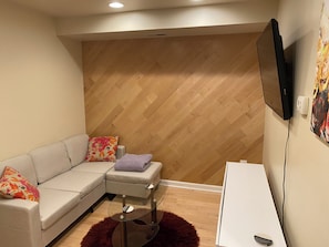 A modern living room with a sectional sofa, a glass coffee table, a small white desk, and a mounted TV.