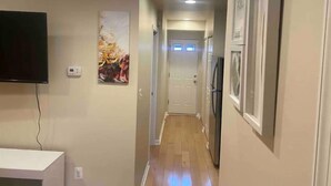A narrow hallway with a flat-screen TV mounted on the left wall, a door at the end, and a refrigerator on the right.