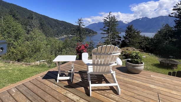 A deck with a white Adirondack chair and a small table, overlooking a lake and mountains.