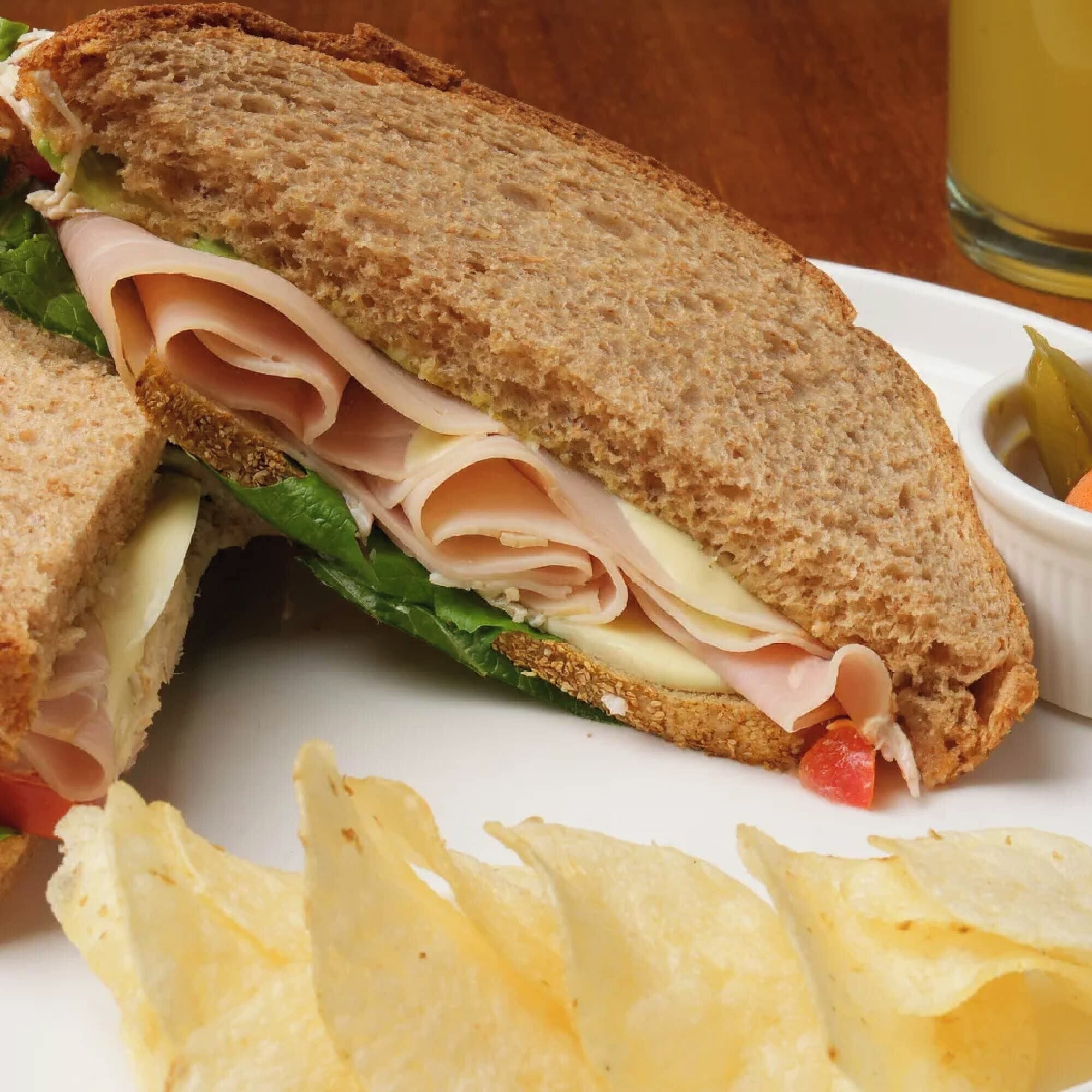 A sandwich with lettuce, cheese, and ham, accompanied by potato chips and a small bowl of pickles.