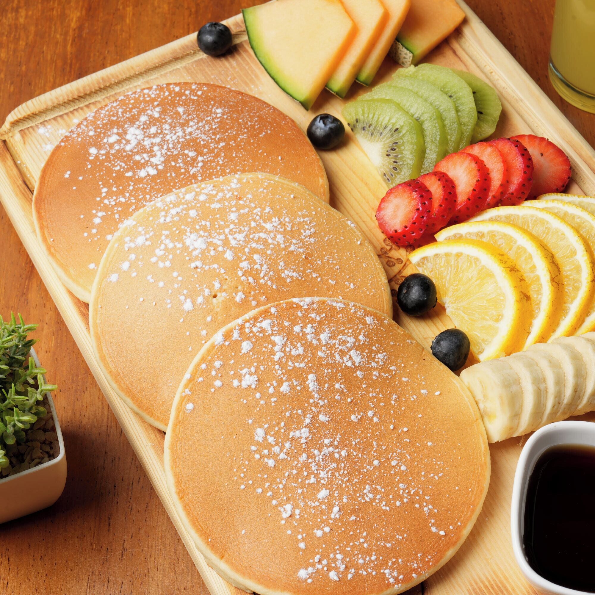 Drei Pfannkuchen auf einer Holzbrett, bestreut mit Puderzucker, begleitet von Obstscheiben und einer kleinen Schüssel Ahornsirup.