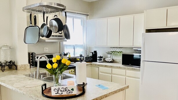 A modern kitchen with white cabinets, a central island, hanging pots, and a black trash bin. - Historic Old Towne Southern Charm (Portsmouth)