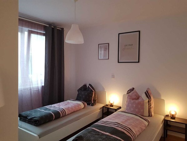 A bedroom with two beds, a window with curtains, a pendant light, and two framed pictures on the wall.