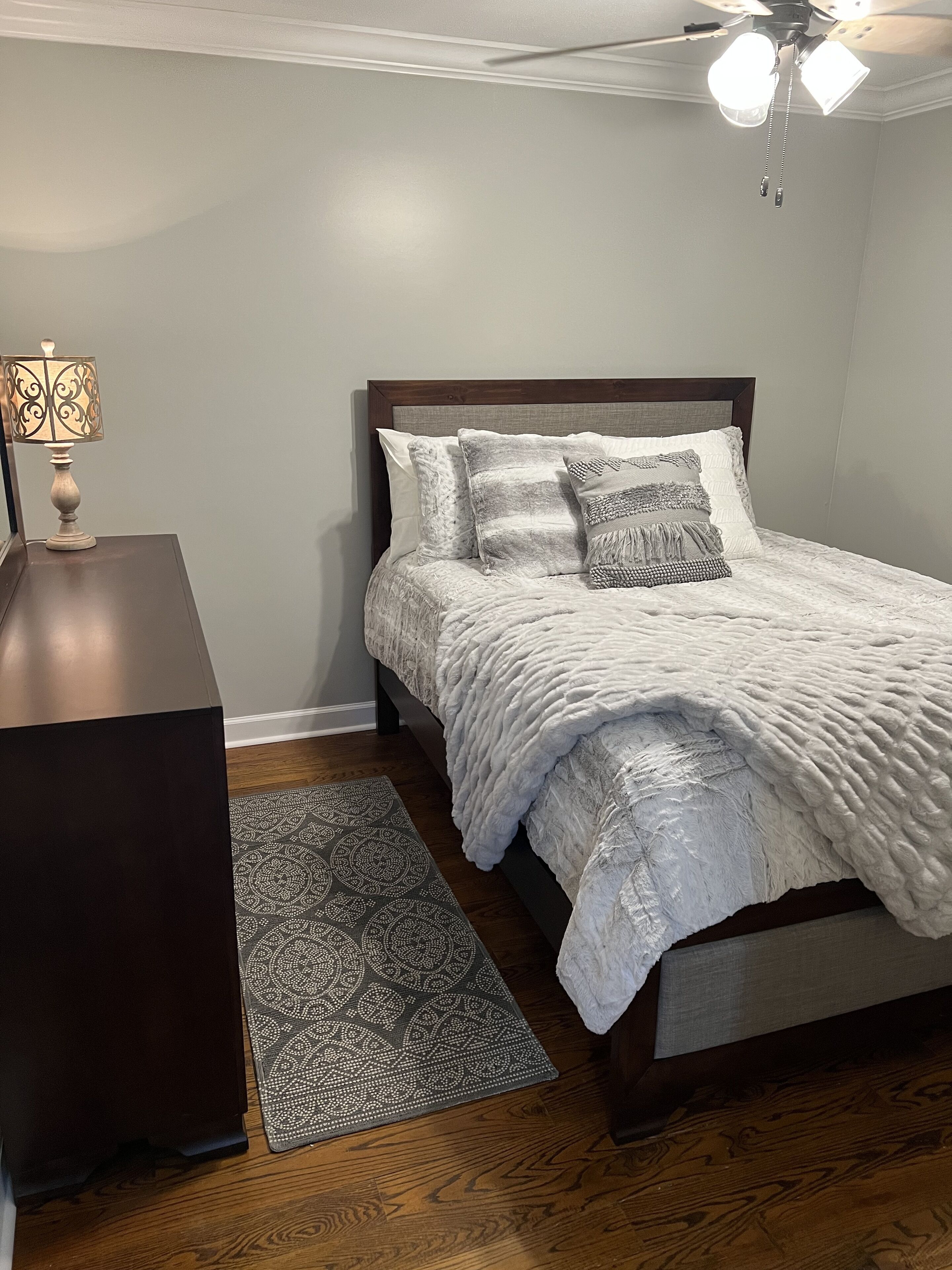 Cama bem arrumada com manta branca texturizada, tapete estampado e mesa de cabeceira com abajur.
