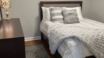 Cama bem arrumada com manta branca texturizada, tapete estampado e mesa de cabeceira com abajur.
